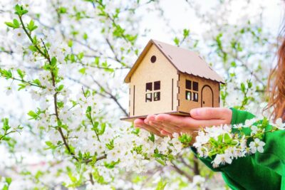 female holds a wooden house model under a blooming tree outdoors. spring buying season concept.